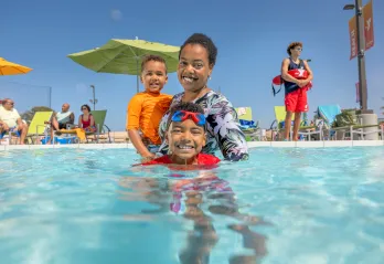 A mother and her toddler and child swim in a YMCA outdoor pool supervised by a YMCA Lifeguard