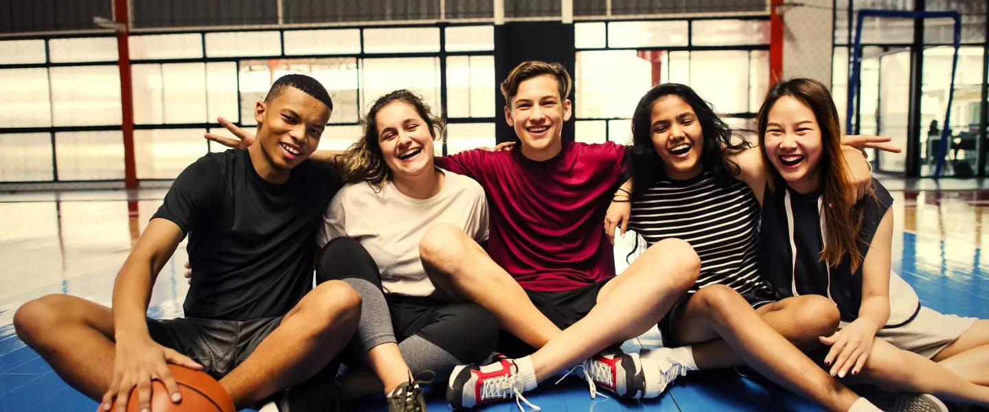 An image of a group of teenagers from the YMCA teen program linked arm-in-arm sitting on a basketball court. From left to right is an African American male, a Caucasian female and male, an African American female, and an Asian female. Everyone is smiling and the male in the middle is making peace signs on his friends shoulders.