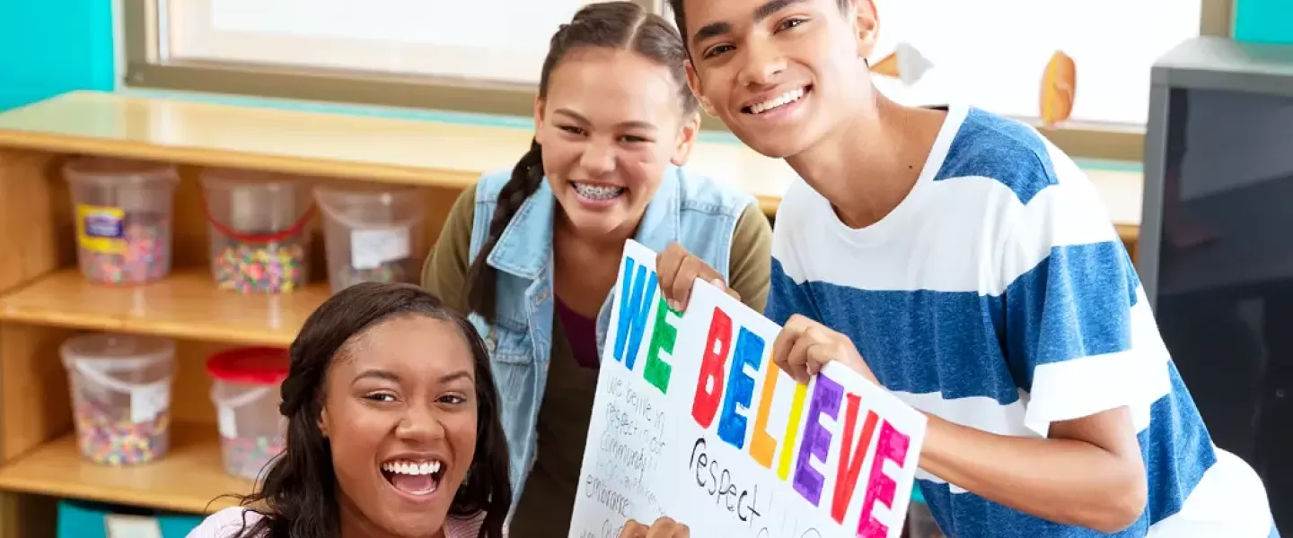 An image of three teens making a colorful sign together that says, "we believe," as well as other words like "respect." The African American female, Asian female, and Middle Eastern male are all looking at the camera, smiling.