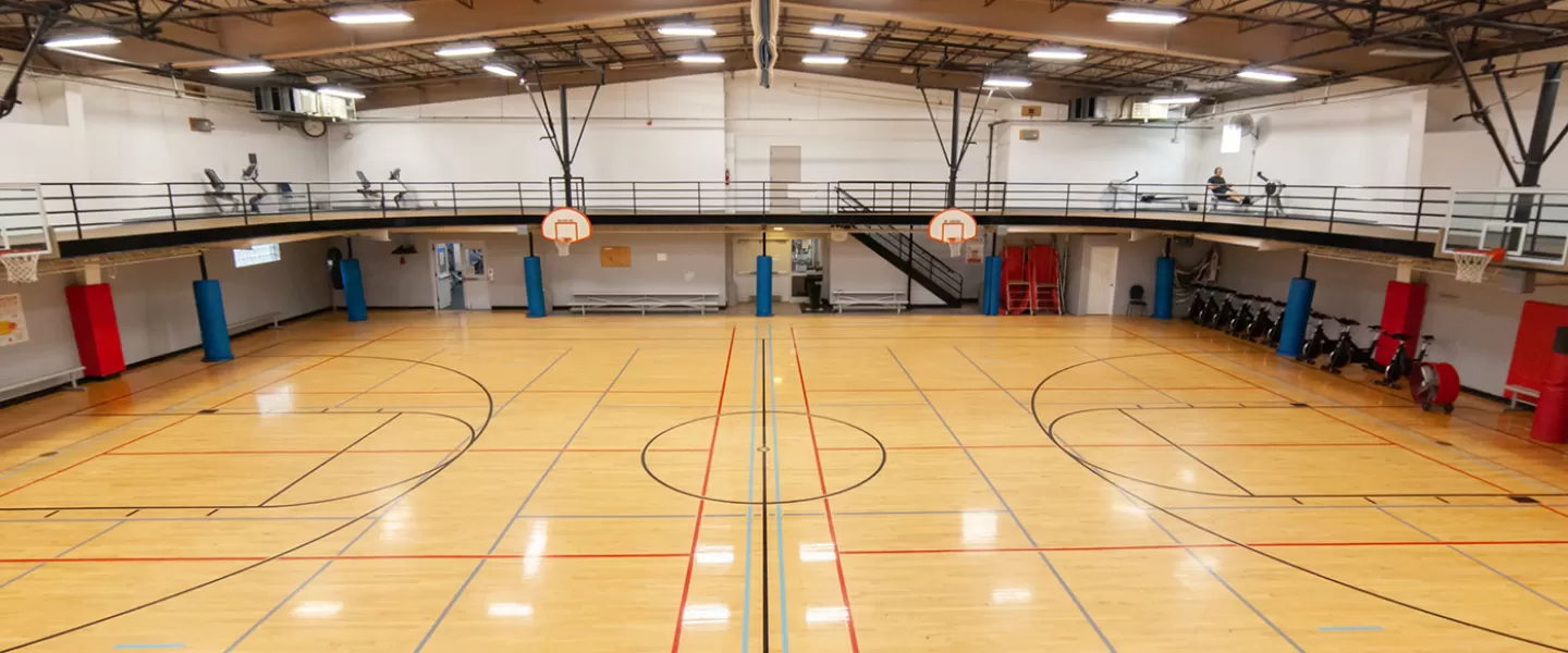 Gymnasium with multiple hoops. Small bench seats along walls of court. Large indoor track running overhead with machines in corners.