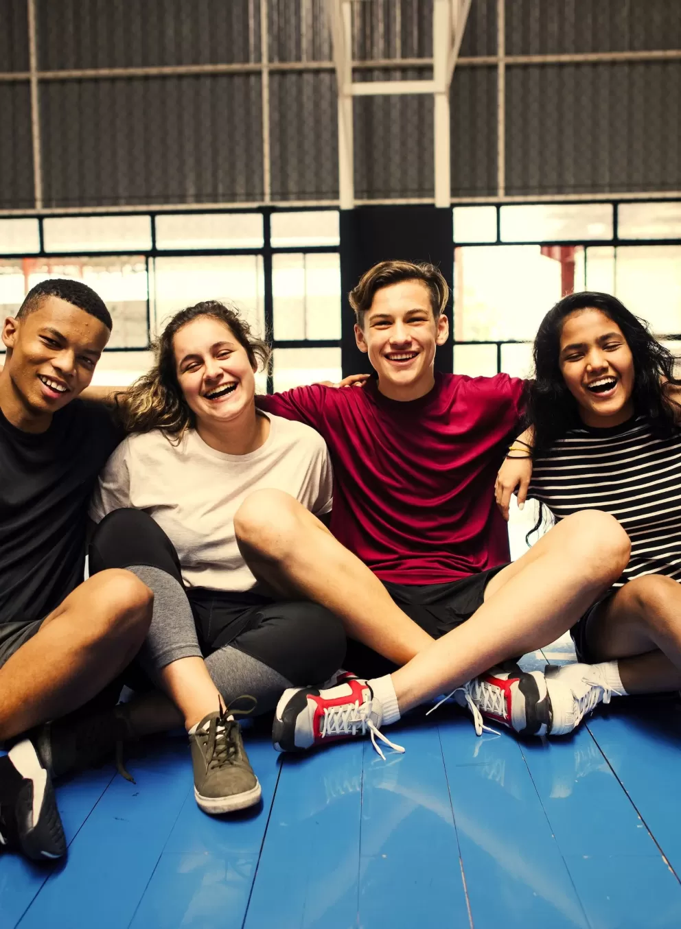 An image of a group of teenagers from the YMCA teen program linked arm-in-arm sitting on a basketball court. From left to right is an African American male, a Caucasian female and male, an African American female, and an Asian female. Everyone is smiling and the male in the middle is making peace signs on his friends shoulders.
