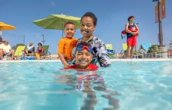 A mother and her toddler and child swim in a YMCA outdoor pool supervised by a YMCA Lifeguard