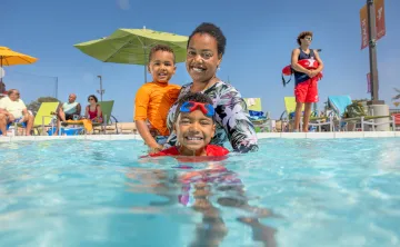 A mother and her toddler and child swim in a YMCA outdoor pool supervised by a YMCA Lifeguard